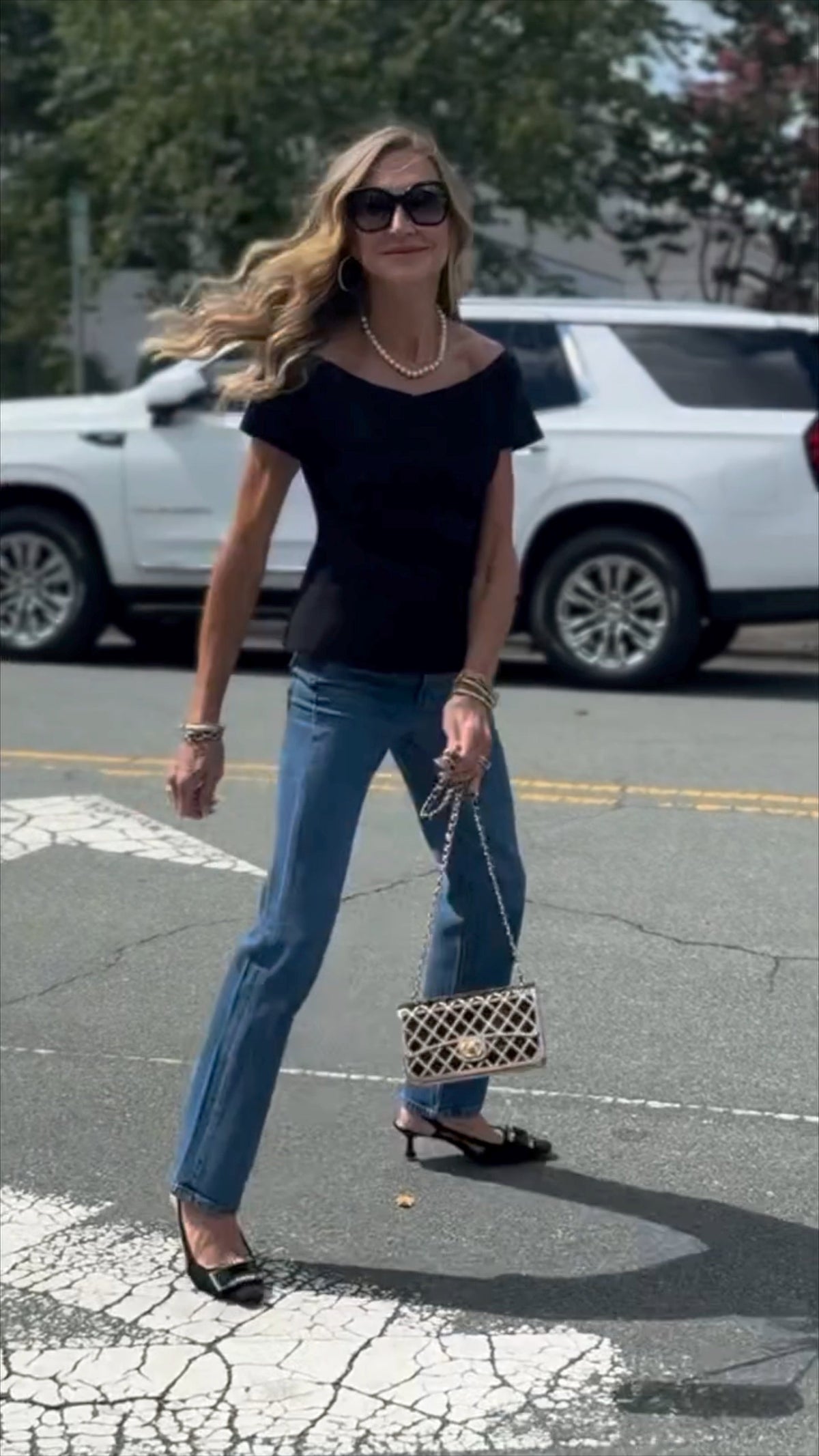 Woman in black top and blue jeans walking on a street with a white car in the background.