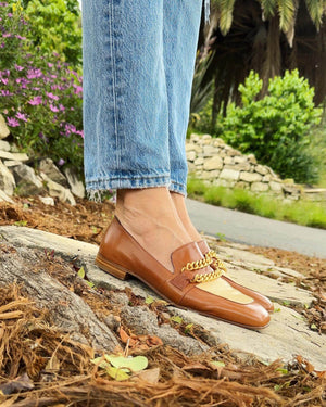 Tan flat loafers on feet with cream inserts and gold chain detail; low heel