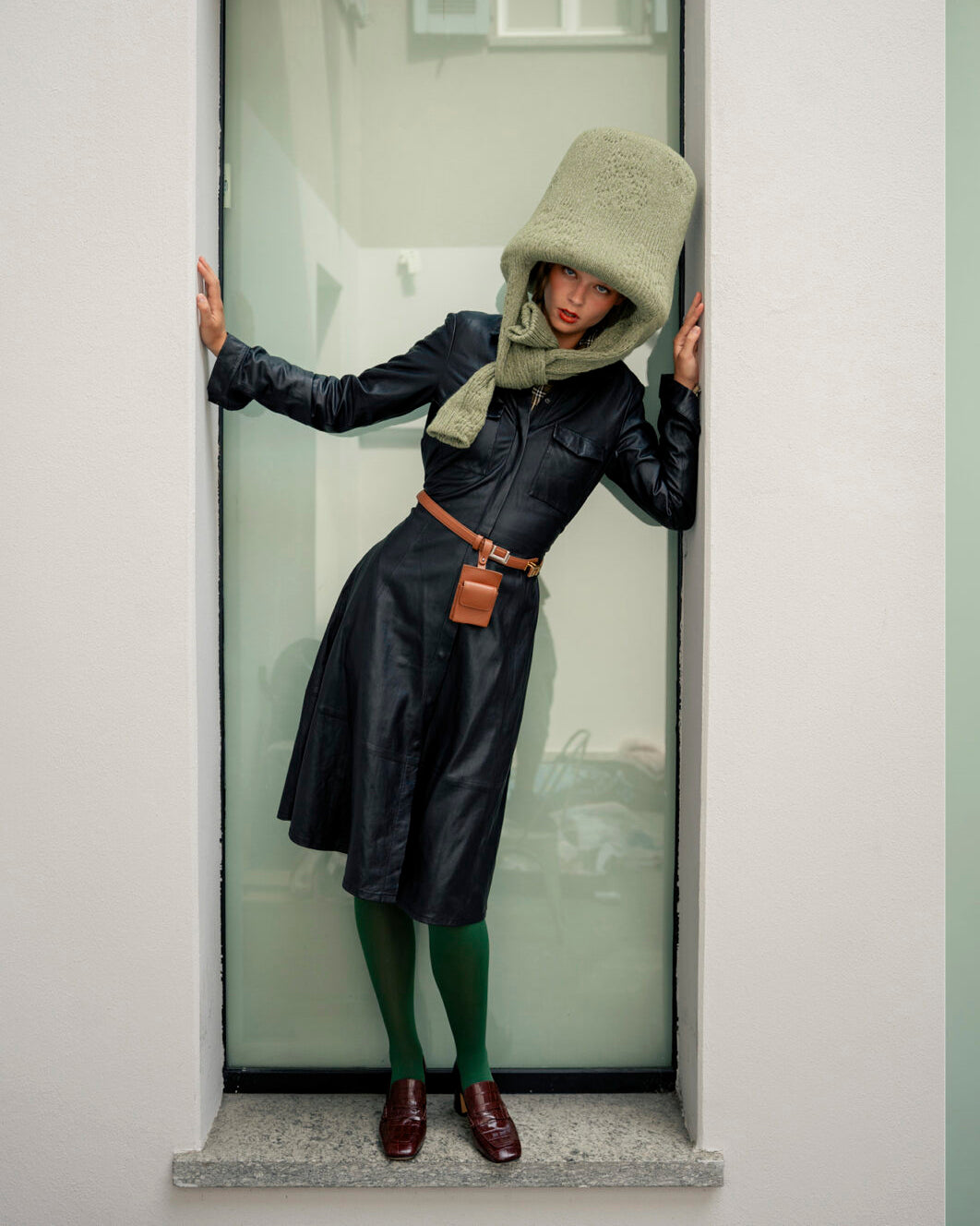 Person wearing a dark coat with a hood, green scarf, and green socks standing inside a glass door.  CAKE Magazine Editorial  featuring Brown Moc Croc Loafers by madison maison