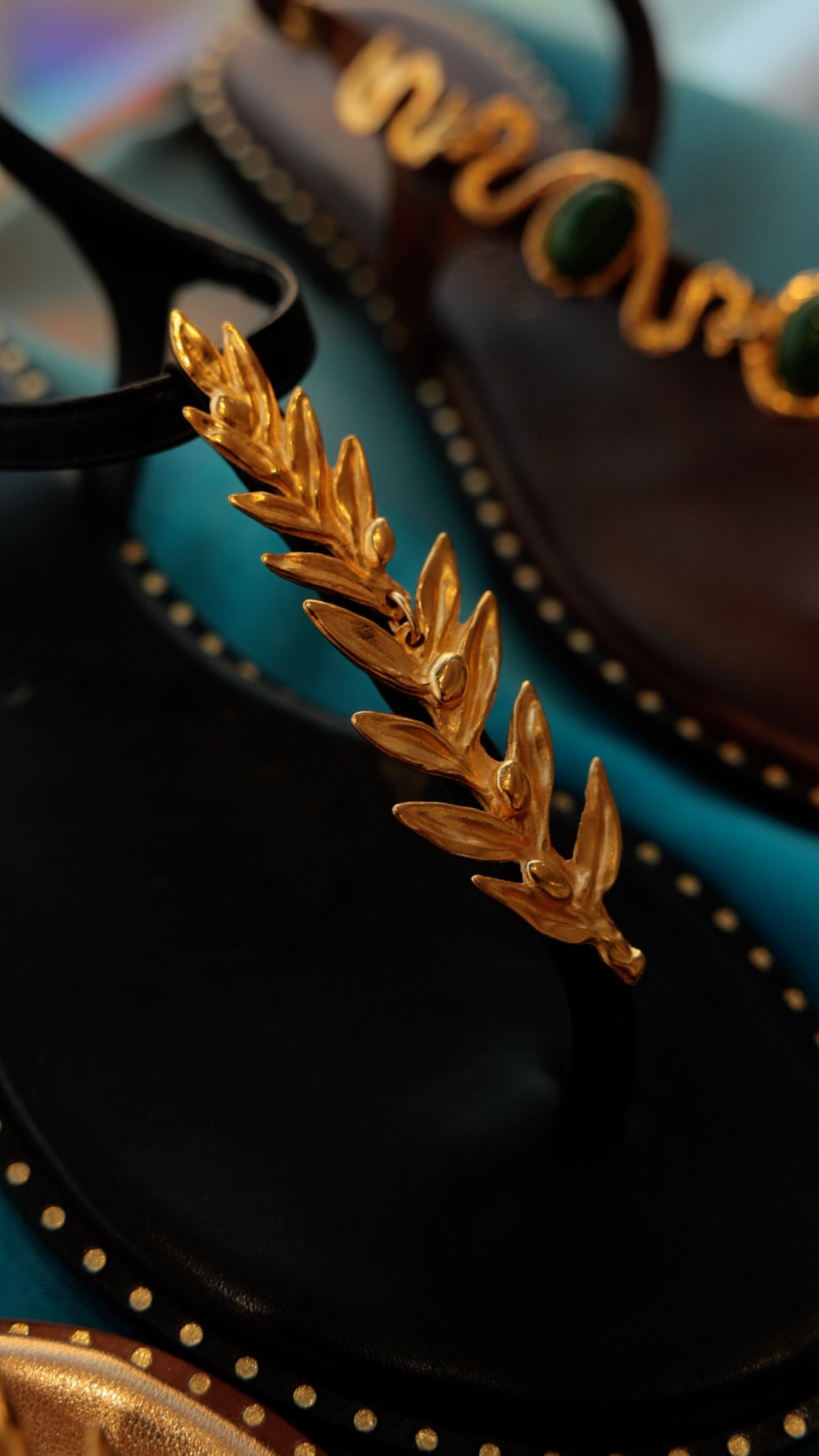 Editorial close-up of the gold leaf detail on a Madison Maison black flat thong sandal with the serpent malachite sandal in soft focus behind.
