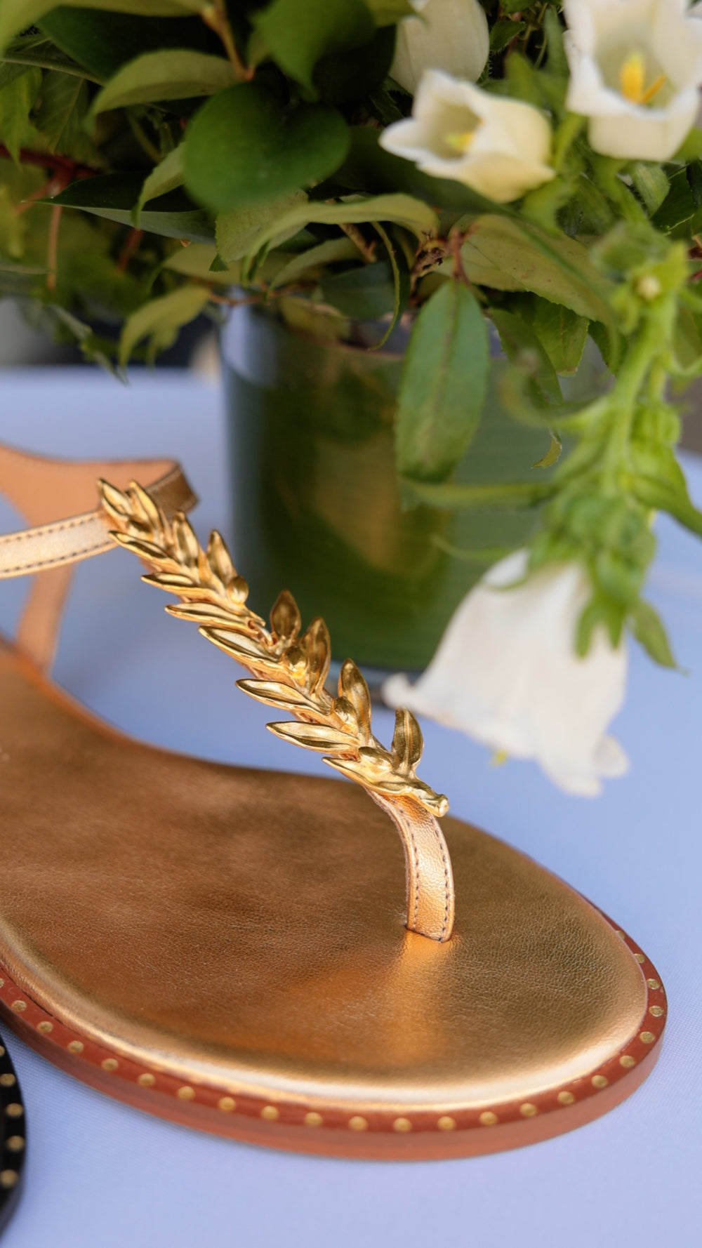 Madison Maison gold metallic leaf thong sandal styled beside white calla lilies and bell flowers in greenery.