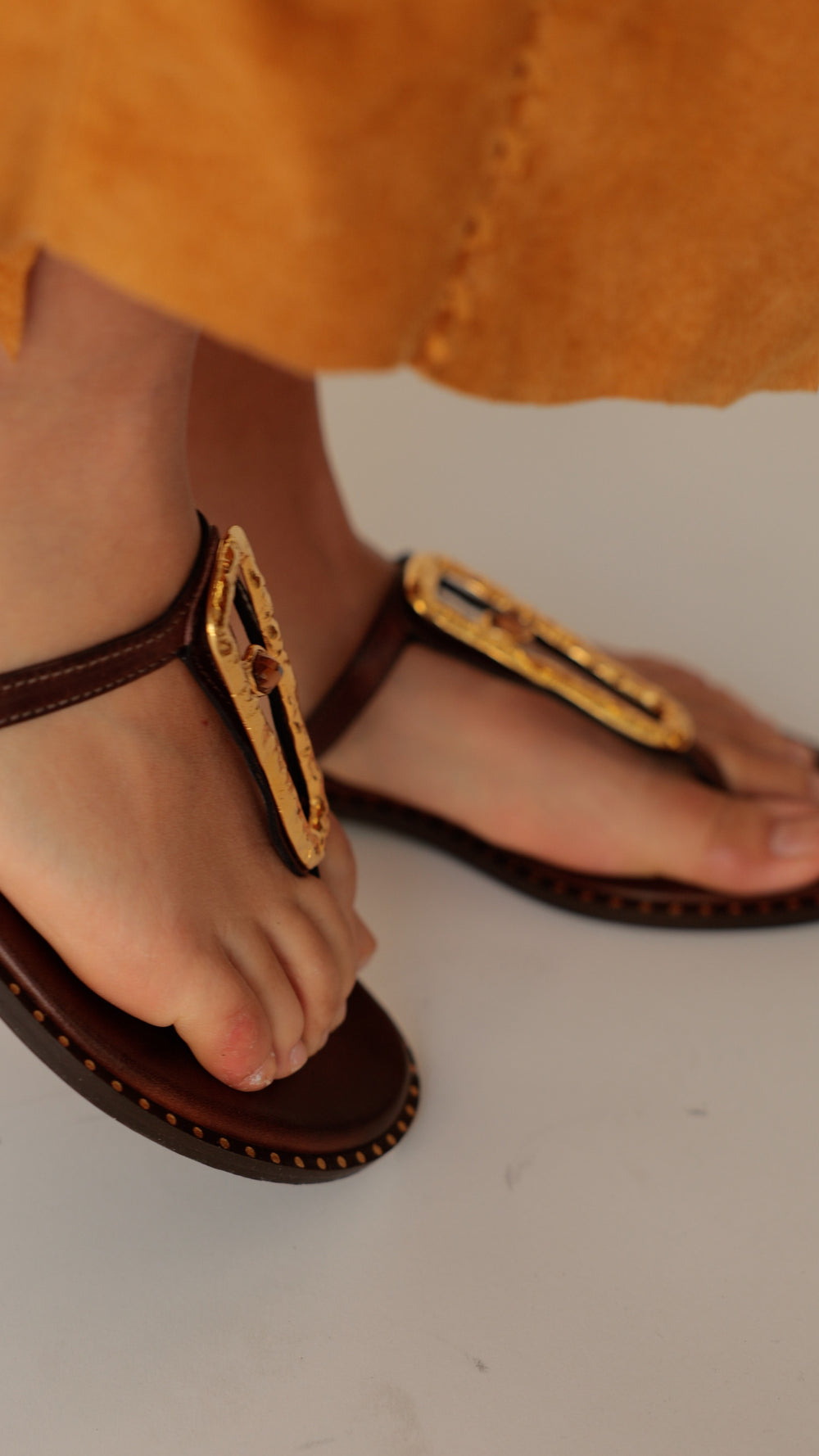 Feet in Madison Maison brown leather hammered hardware flat sandals, styled with a saffron suede scalloped skirt.