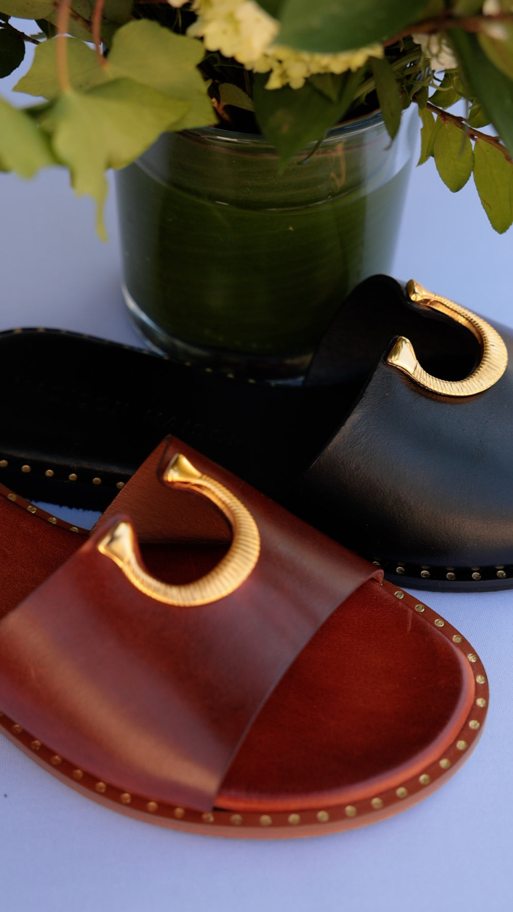 Madison Maison horse shoe buckle leather flat sandals in black and caramel with brass studded soles, styled beside hydrangeas in a green vase.