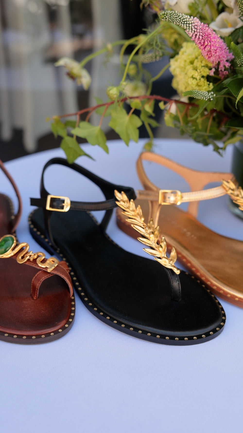Three Madison Maison flat sandals together: gold leaf thong on black, chocolate serpent with green stone, and gold metallic leaf thong, with a fresh floral arrangement.