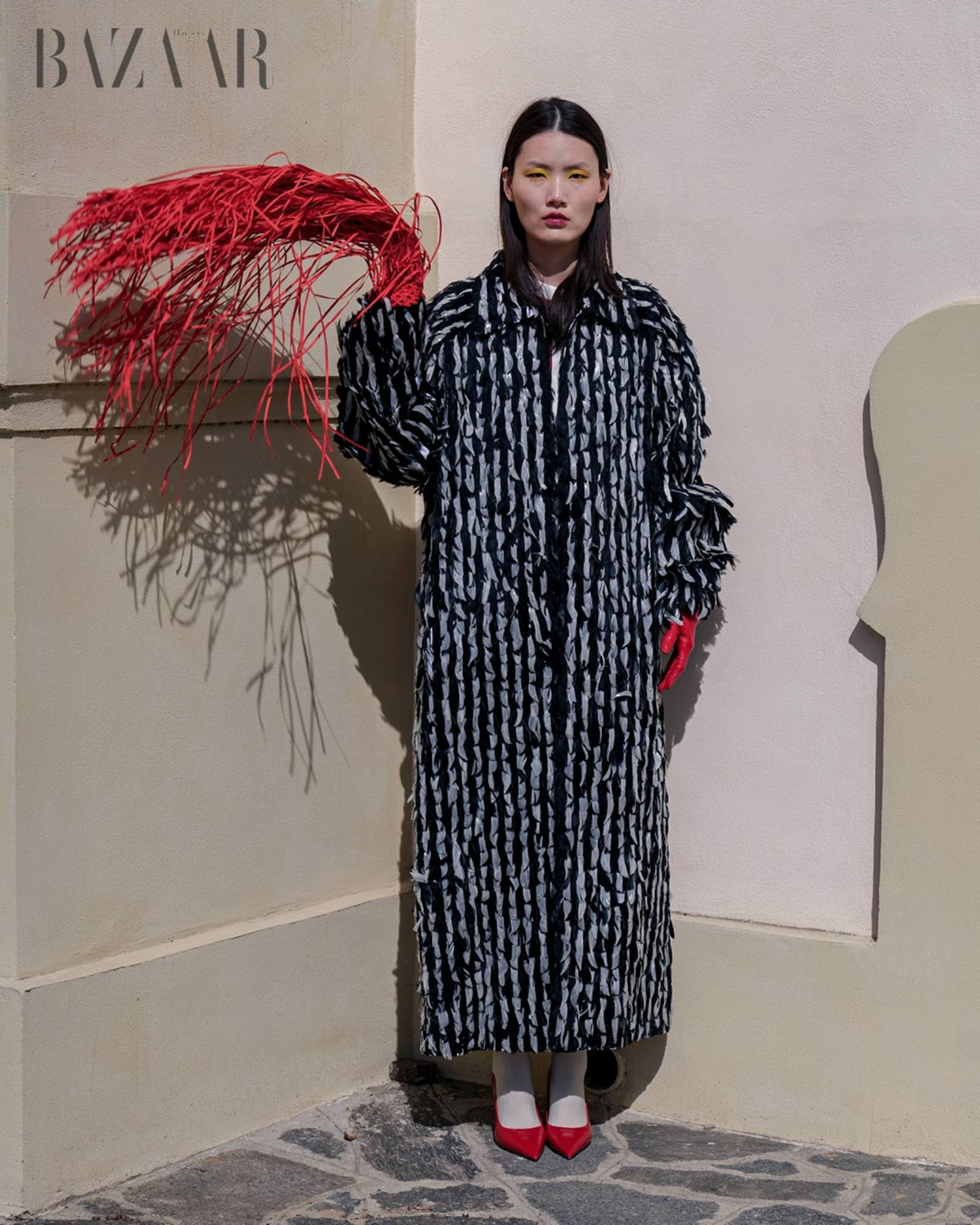 Model wearing a black and white patterned coat with red accents, standing next to a decorative red plant against a beige wall.